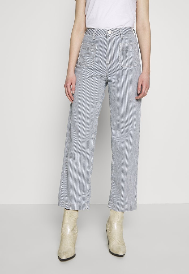 Femme portant un pantalon taille haute, à jambes larges, rayé bleu clair et blanc avec poches avant, un haut blanc sans manches rentré, et des bottines crème.