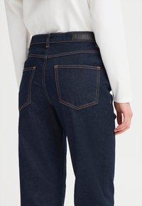 Persona que lleva puestos unos vaqueros de mezclilla oscuros con costuras naranjas contrastantes y una camiseta blanca de manga larga, vista posterior mostrando los bolsillos y la etiqueta de la cinturilla.