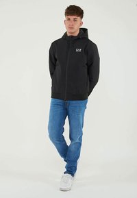 Jaunas vyras, vilkintis juoda EA7 zip-up gobtuvu striuke, mėlynomis džinsomis ir baltomis sportinėmis bateliais, stovi su rankomis už nugaros.