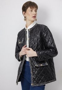 Black quilted jacket with a textured finish, trimmed in light contrast thread, featuring two patch pockets and gold-tone buttons.