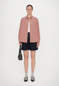 Chemise rose clair à manches longues, avec poche avant, associée à un short en jean foncé, des chaussettes grises et des chaussures noires. Modèle tenant un sac à main noir.