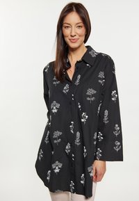Camicia nera con bottoni, ricamo floreale bianco, maniche lunghe e vestibilità rilassata. Presenta un colletto e una tasca sul lato sinistro del petto.