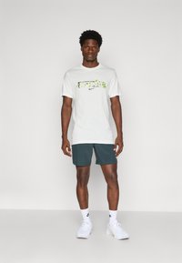 Biela bavlnená tričko s zelenou grafikou "JUST DO IT" a logom Nike, spárované s tmavozelenými šortkami a bielymi športovými topánkami.