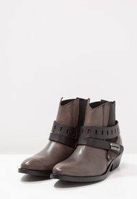 Brązowe skórzane botki za kostkę z wygiętym obcasem, elastycznymi panelami bocznymi oraz czarnym paskiem z wyciętymi wzorami i srebrną klamrą jako akcentem.