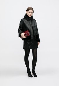 Schwarze Lederjacke mit Pelzkragen, über einem schwarzen, strukturierten Blumenkleid getragen. Hält eine burgunderrote Handtasche, trägt schwarze Strumpfhosen und Schuhe.