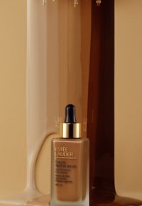 Sérum teinte de peau Futurist d'Estée Lauder dans un flacon en verre transparent avec un compte-gouttes noir, présentant un capuchon et une étiquette dorés, sur un fond neutre.