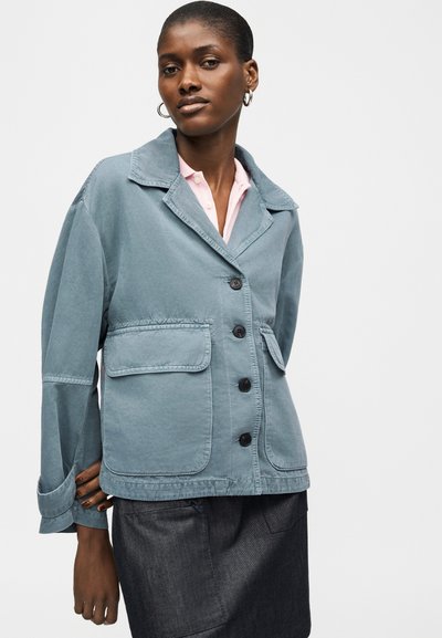 Femme portant une veste bleue boutonnée avec de grandes poches avant et une jupe sombre, debout devant un fond clair uni.
