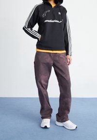 Black zip-up hoodie with white logo and stars, paired with orange shirt and dark marbled pants. White athletic shoes complete the look.