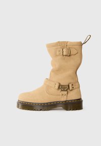 Kniehoge beige suède laarzen met een gespsluiting en een gegraveerd logo, voorzien van een dikke, gestructureerde rubberen zool met gele stiksels.