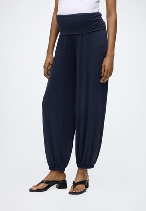 Personne portant un pantalon bleu marine ample avec des poignets élastiques et des sandales à talons noires, debout sur un fond blanc.