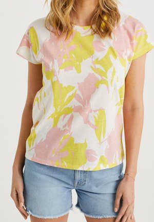 Kobieta w pastelowo żółtej i różowej koszulce w kwiatowy wzór oraz jasnoniebieskich szortach jeansowych, stojąca z rozluźnionymi ramionami.