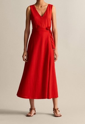 Sleeveless red midi dress with V-neck and side tie belt, worn with brown ankle-strap sandals on a neutral background.