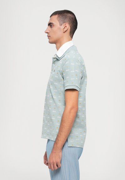 Cămașă polo bleu deschis cu guler alb, având un design cu modele de bărci mici și accente aurii. Purtați cu pantaloni de înălțime deschisă cu dungi.
