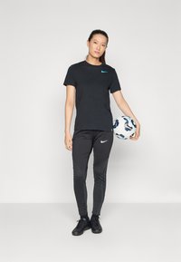 Musta Nike t-paita, jossa on turkoosi logo, mustat Nike housut, joissa on teksturoidut yksityiskohdat, pitää kädessään kuvioitua jalkapalloa, seisoo yksinkertaisella taustalla.