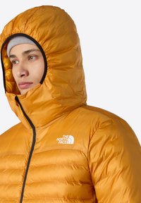 Přefouknutá bunda v živé oranžové barvě s prošívaným designem a vysokým límcem. Obsahuje černý zip a malé bílé logo na prsou.