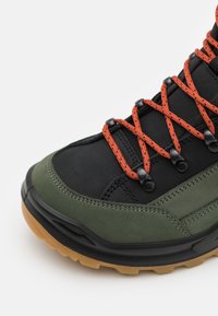 Schwarzer und grüner Wanderschuh mit orangenen Schnürsenkeln, Gummisohle, strukturierten Einsätzen und Metallösen für die Schnürsenkel. Strapazierfähiges synthetisches Material.