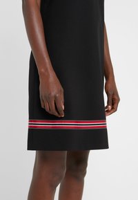 Robe noire avec des rayures horizontales rouges, blanches et noires près de l'ourlet, portée par une personne à la peau foncée, visible de la mi-cuisse à la taille.