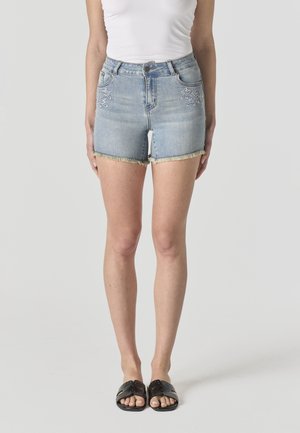 Mujer que lleva shorts vaqueros bordados en azul claro con dobladillos deshilachados, top sin mangas blanco y sandalias negras sin cierre con tachuelas.