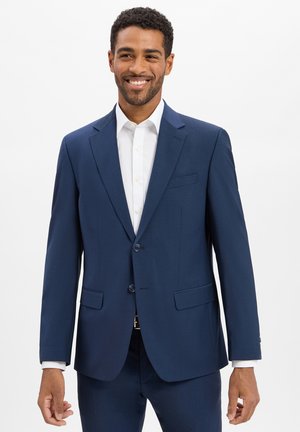 Smiling man wearing a dark blue suit jacket and matching pants over a white button-up shirt against a plain white background.