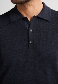 Marineblauwe poloshirt gemaakt van een zachte, getextureerde knitstof. Bevat een kraagrand en drie zwarte knopen bij de sluiting.