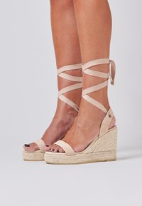 Sandales compensées en espadrille beiges avec des attaches autour de la cheville, présentant une tige lisse, une semelle intermédiaire en jute tissé et un design à bout ouvert.
