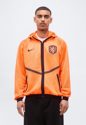 Man draagt een feloranje mesh jas met ritssluiting, zwarte bies en leeuwembleem, staand tegen een effen lichte achtergrond.