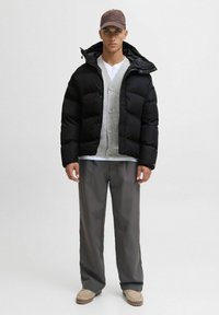 Giacca a vento nera con texture trapuntata, cardigan grigio sotto, camicia chiara, pantaloni scuri e scarpe beige. Accenti includono un berretto.