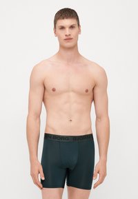 Moške temno zelene bokserice z črnim elastičnim pasom, na katerem je natisnjen naziv blagovne znamke "BJÖRN BORG". Gladka, raztegljiva tkanina.