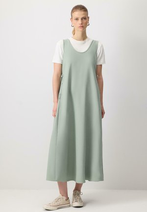 Robe longue sans manches en tissu vert doux avec un col arrondi et des liens sur les côtés, superposée sur un t-shirt blanc et associée à des baskets beiges.