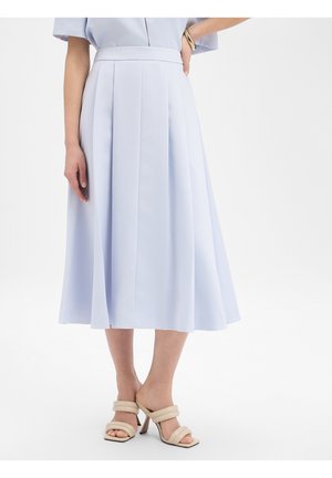 Frau trägt einen hellblauen Midirock mit Falten, kombiniert mit beigen Sandalen mit Absatz, steht mit einer Hand in der Hüfte.