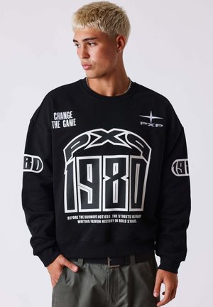 Young man with short blond hair wearing a black sweatshirt with white graphics and text including "1980" and "CHANGE THE GAME."