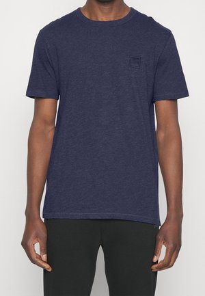 Dunkelblaues T-Shirt mit kurzen Ärmeln und Rundhalsausschnitt an einer Person mit dunkler Hose, mit einem kleinen quadratischen Logo auf der linken Brust.