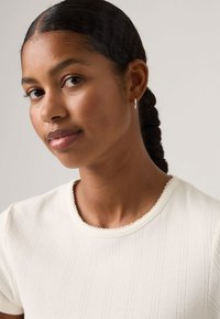 Νεαρή γυναίκα με πλεξούδες, που φοράει μικρούς κρίκους στα αυτιά και λευκό υφασμάτινο κοντομάνικο τοπ, κοιτάζοντας ελαφρώς προς το πλάι.