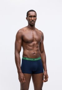 Uomo senza maglietta con corporatura muscolosa che indossa boxer blu navy con una vita verde recante il logo bianco "GANT".