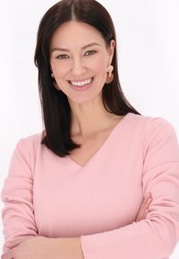 Pulover roz deschis cu decolteu în V și mâneci lungi. Modelul poartă cercei mari, rotunzi, în tonuri de bej, zâmbind pe un fond alb.