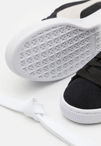 Baskets noires avec une semelle blanche montrant un motif de bande de roulement texturé, des lacets noirs et un lacet blanc noué en nœud à côté sur une surface blanche.