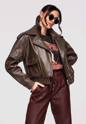 Frau trägt braune, kurz geschnittene Lederjacke, bedrucktes T-Shirt, bordeauxrote Lederhose, schwarze Sonnenbrille und goldene Creolen, posiert mit Händen in den Taschen.