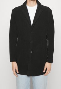 Manteau en laine noire mélangée avec un col cranté, fermeture à bouton sur le devant et deux poches latérales. Texture lisse, coupe ajustée et design minimaliste.