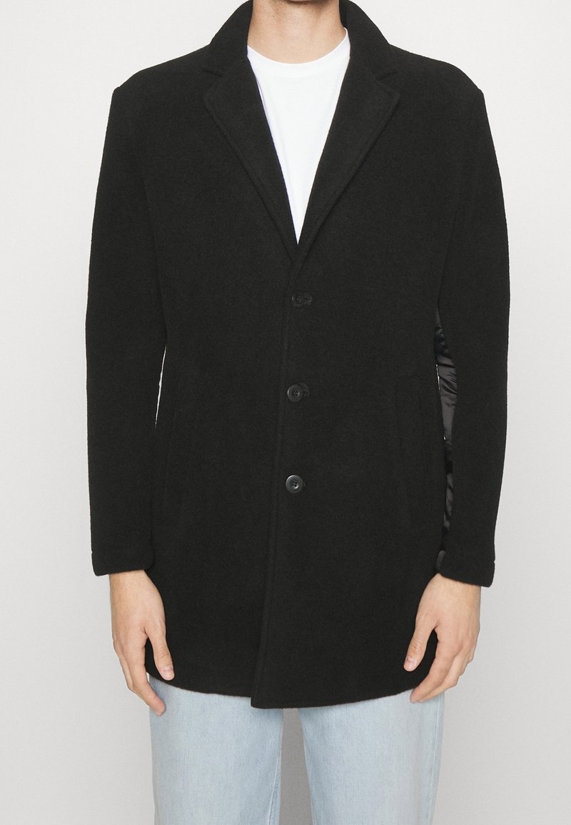 Manteau en laine noire mélangée avec un col cranté, fermeture à bouton sur le devant et deux poches latérales. Texture lisse, coupe ajustée et design minimaliste.