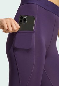 Ljubičaste helanke visokog struka s bočnim džepom za pametni telefon. Izrađene od glatkog, rastezljivog materijala s vidljivim šavovima.