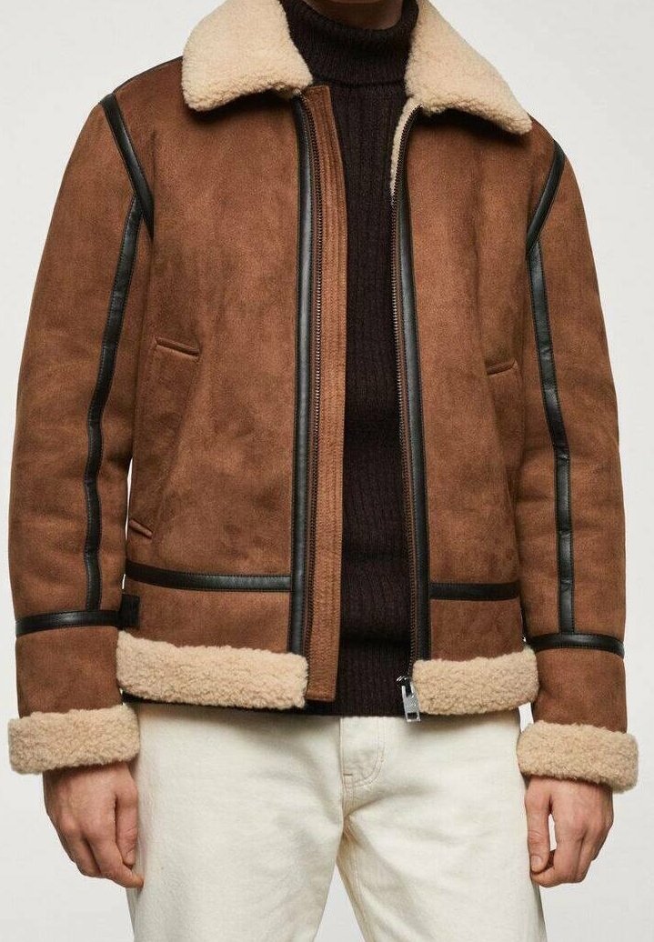 Bruine shearling jas met een crèmekleurige fleece kraag en manchetten, zwart leren detail, gedragen over een donkerbruine trui en witte broek.