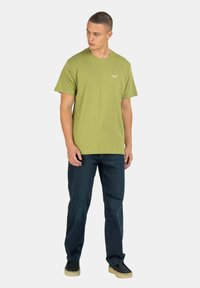 Camiseta de algodón verde oliva con mangas cortas y cuello redondo. Presenta un discreto bordado del logo en el pecho. Se lleva con jeans oscuros y zapatos negros.