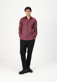 Chemise boutonnée bordeaux à manches longues, associée à un pantalon noir et des chaussures noires. La chemise présente une texture subtile et une coupe classique.