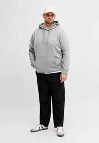 Grauer Kapuzenpullover mit Fronttasche und Kordelzug, versehen mit dem "JACK & JONES"-Logo. Getragen mit schwarzen Hosen und beigefarbener Mütze, kombiniert mit Turnschuhen.