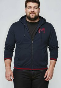 Felpa zip con cappuccio color navy, con coulisse, righe rosse sui polsini e sull'orlo, e logo bordeaux sul petto. Texture del tessuto liscia.