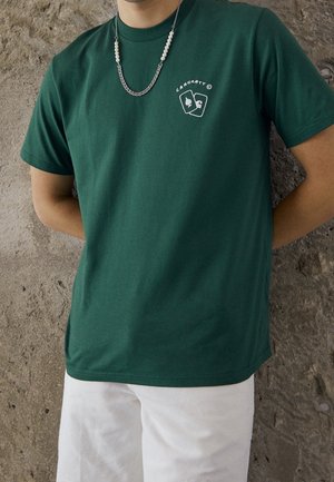 Persoon draagt een donker groen Carhartt t-shirt met klein logo op de borst en gelaagde zilveren en parelkettingen, staand tegen een gestructureerde stenen muur.