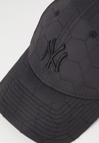 Black cap with hexagonal quilted texture, featuring an embroidered logo in black. Soft fabric with a curved brim and stitched accents.