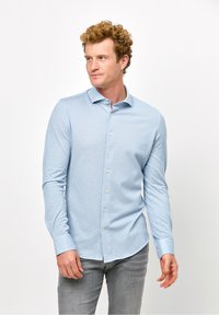 Camicia azzurro chiaro a maniche lunghe con colletto button-down, caratterizzata da una texture liscia, bottoni frontali e un orlo curvo. Abbinata a pantaloni grigi.