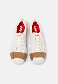 White leather sneakers with a brown rubber toe cap,  lace-up closure, and red interior featuring "CAMPER" branding. Smooth texture and modern design.