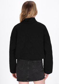 Sort quiltet jakke med høj krave, der har en afslappet pasform. Bæres over en sort denimnederdel med baglommer. Teksturen ser blød ud.
